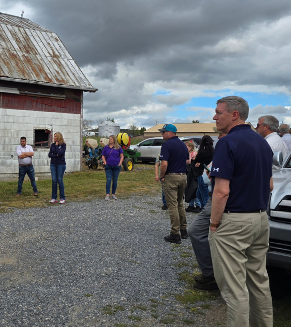 MD Agriculture Secretary Kevin Atticks visits Garrett County farms. Photo of Secretary Atticks and other individuals before a background of a barn and sky with dark clouds.