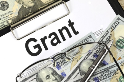 The word "Grant" in black text on a background of a white clipboard, paper U.S. money, a pen and glasses.