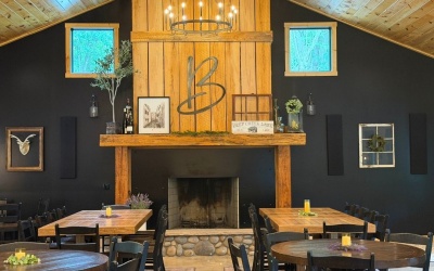 Interior of Blackthorne Cellars featuring a rustic stone fireplace, large wooden mantel with a decorative “B” emblem, wood-paneled ceiling and walls, chandelier, and tables arranged throughout the dining area.