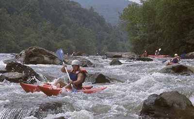 Photo: A kayaker wearing a helmet and life jacket paddles through whitewater rapids on a rocky river, with several other kayakers navigating the rapids in the background and forested hills surrounding the river.