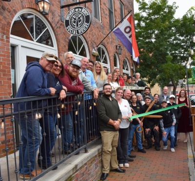 Community and business leaders gather outside Brewery and the Beast Social House in downtown Oakland for a ribbon-cutting ceremony. The group stands in front of the brick building as business owner Craig Frazee and others hold a green ceremonial ribbon and oversized scissors.