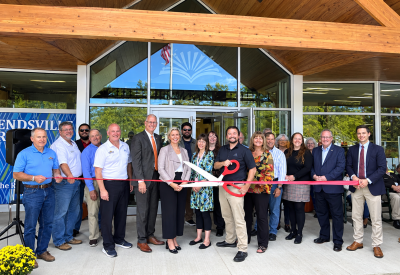 Community members, local officials, and library representatives gather for a ribbon-cutting ceremony outside the new Friendsville branch of the Ruth Enlow Library in Garrett County, Maryland. Participants hold oversized scissors and a red ribbon in front of the building’s glass entrance. Photo courtesy of Claudia Reynolds, The Garrett County Republican.